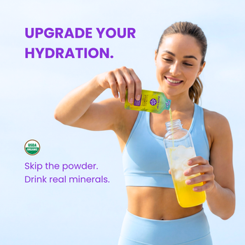 A woman in a light blue sports bra and matching shorts pours a yellow beverage from a green juice box into a clear water bottle with ice. Text on the image reads 'UPGRADE YOUR HYDRATION.
